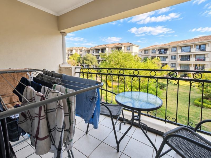 A covered balcony featuring tiled flooring and a decorative wrought iron balustrade, overlooking a landscaped garden and surrounding apartment complex.