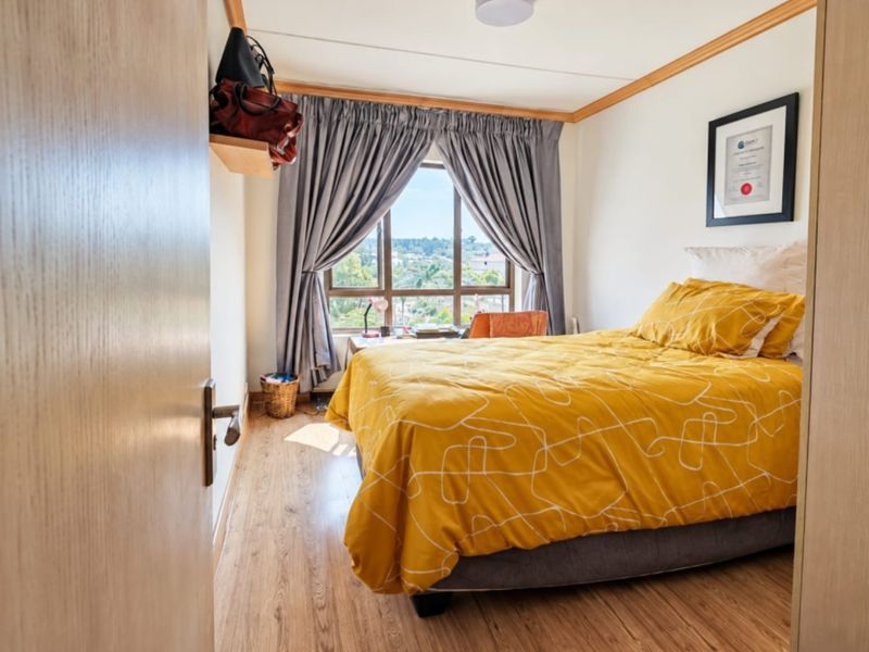 A well-lit bedroom featuring laminate flooring, a large window with natural light, and neutral wall finishes.