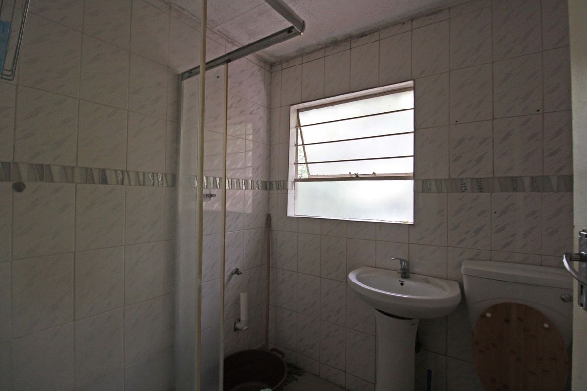 A dated bathroom featuring a pedestal basin, toilet, and shower enclosure with full-height wall tiling and a louvered window.