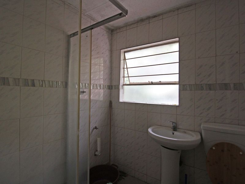 A dated bathroom featuring a pedestal basin, toilet, and shower enclosure with full-height wall tiling and a louvered window.