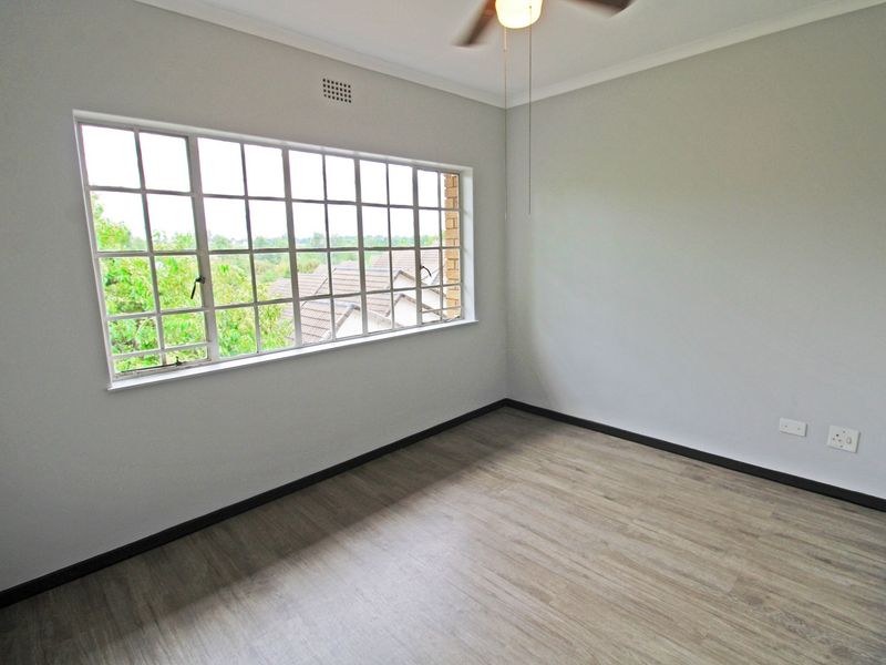 A bright, empty bedroom featuring modern laminate flooring, a ceiling fan, and a large grid-pattern window providing natural light.