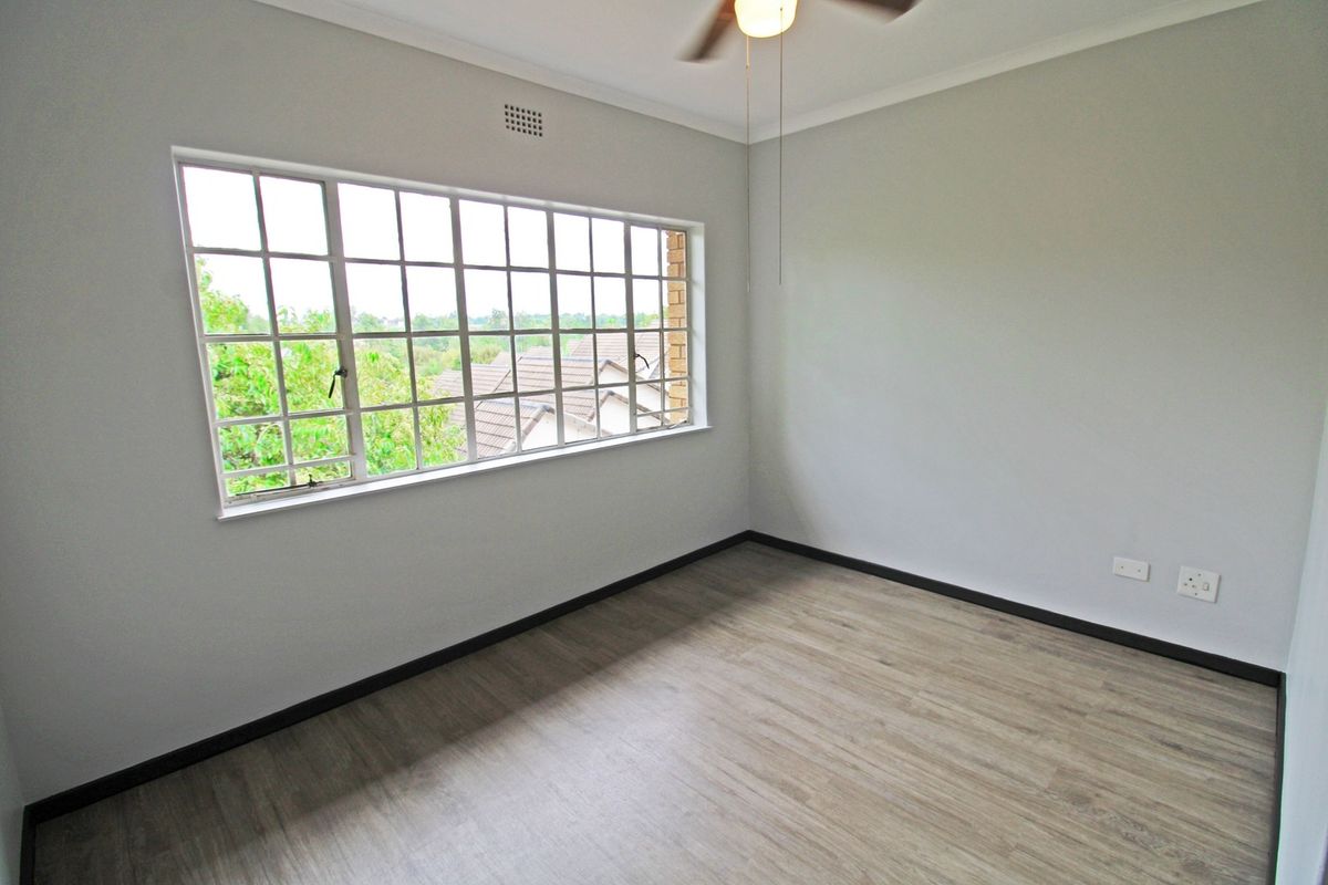 A bright, empty bedroom featuring modern laminate flooring, a ceiling fan, and a large grid-pattern window providing natural light.