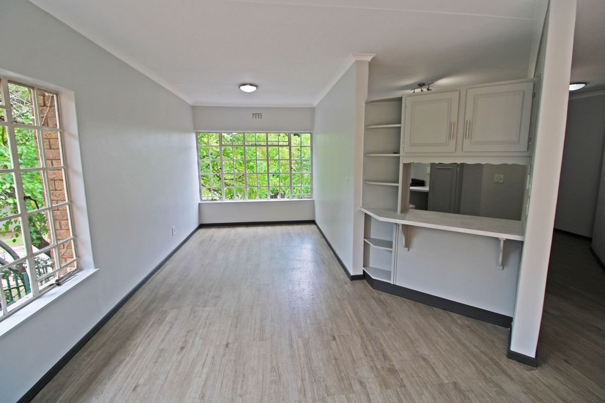 An empty living area featuring light laminate flooring, built-in cabinetry with a breakfast bar, and large steel-framed windows overlooking greenery.