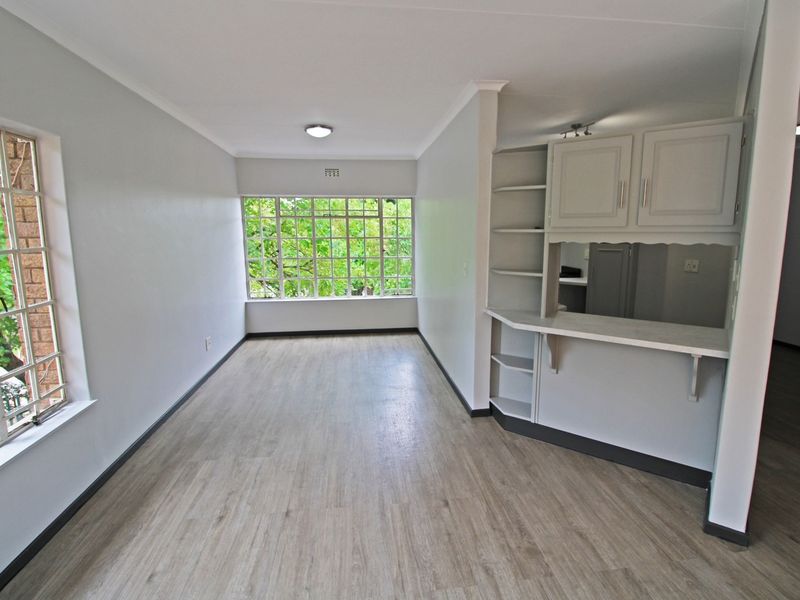 An empty living area featuring light laminate flooring, built-in cabinetry with a breakfast bar, and large steel-framed windows overlooking greenery.