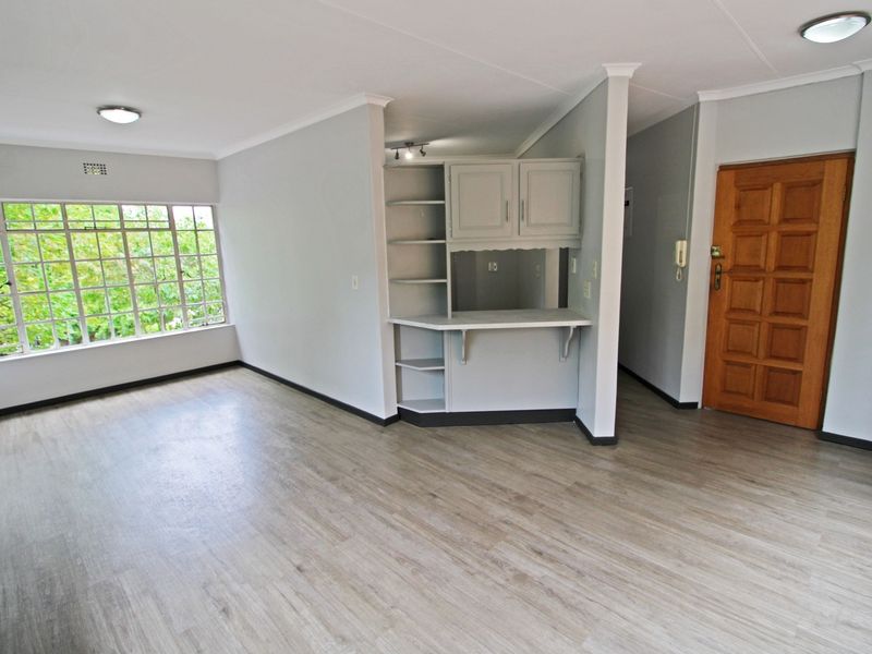 An open-plan living area featuring light-toned laminate flooring, neutral walls, built-in cabinetry, and a large window providing natural light.