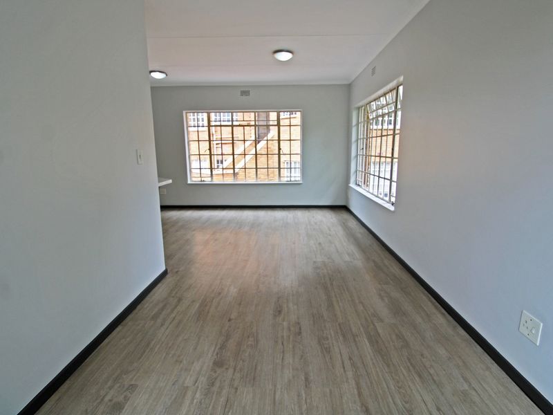 A bright, empty living area featuring modern laminate flooring, neutral walls, and large steel-framed windows providing ample natural light.