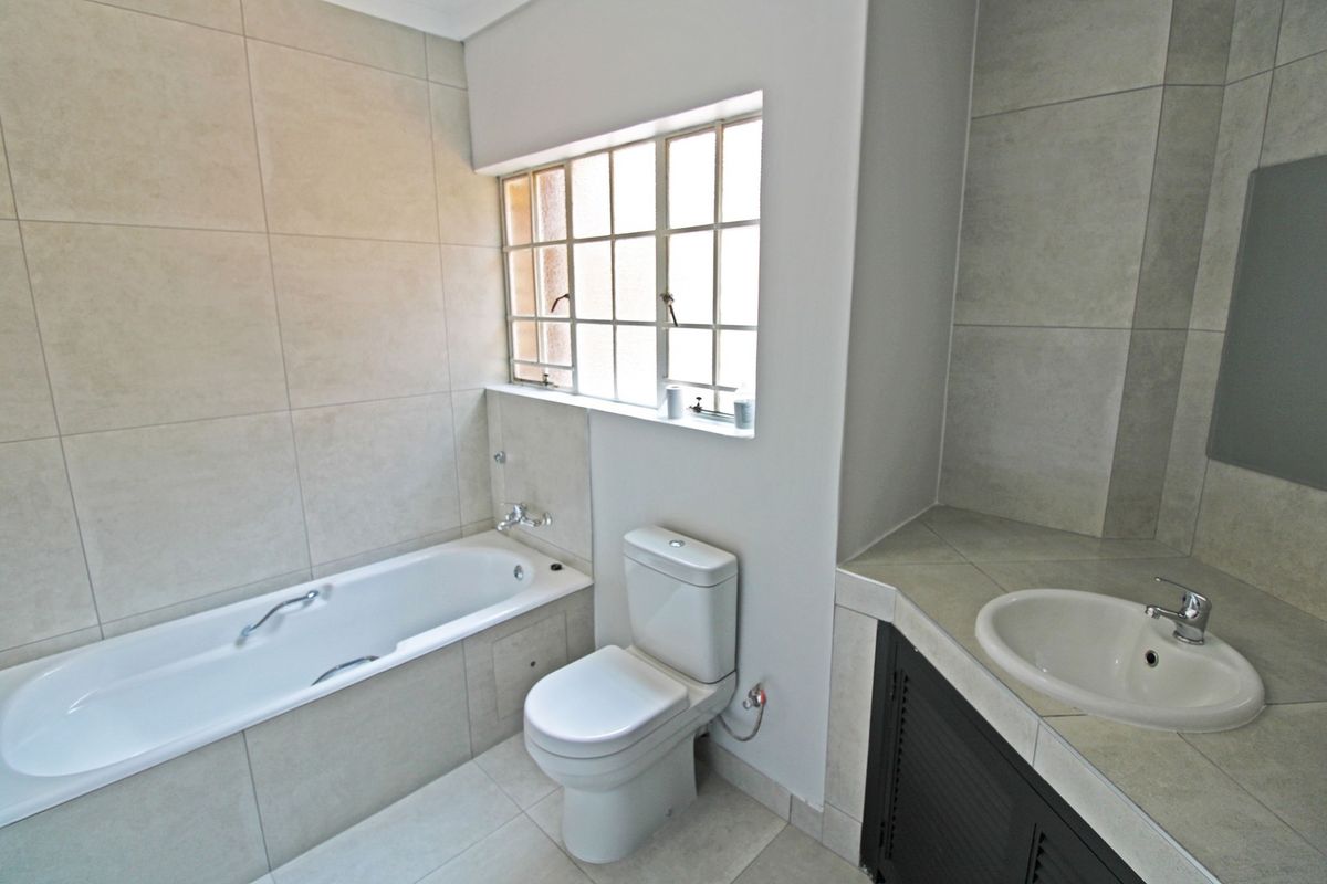 A clean, neutral-toned bathroom featuring a built-in bathtub, a modern toilet, and a vanity basin with tiled finishes throughout.