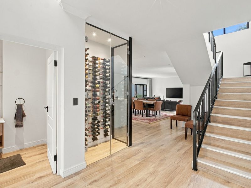 A bright hallway featuring a modern glass-enclosed wine cellar, a staircase with a sleek metal balustrade, and light wood-look laminate flooring.