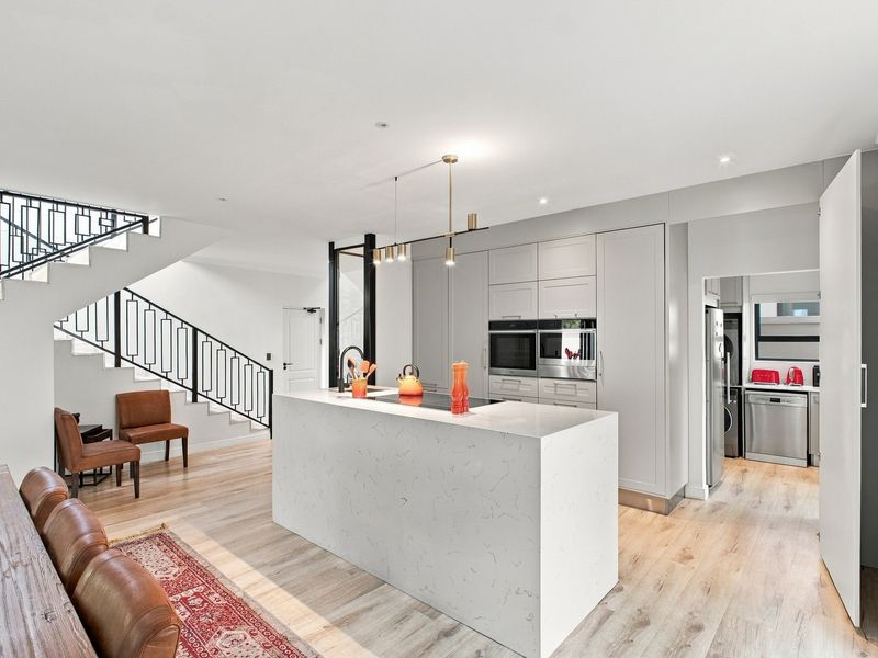 Modern open-plan kitchen featuring a large stone-topped island, integrated appliances, and a sleek staircase, seamlessly connecting to a functional scullery area.