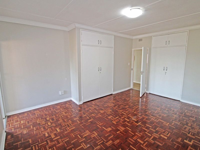 A spacious, empty bedroom featuring classic parquet flooring, neutral walls, and ample built-in cupboard storage with overhead cabinets.
