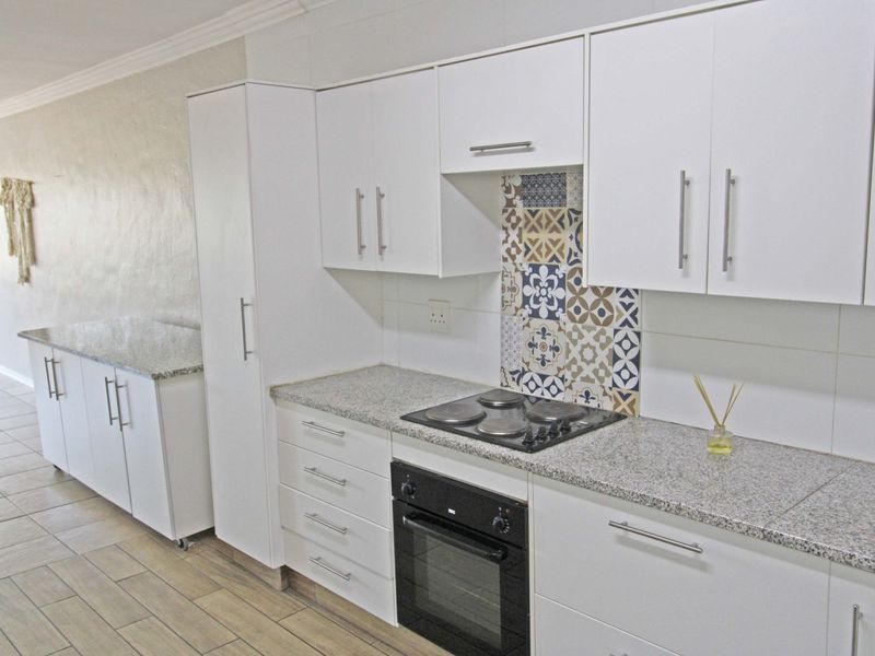 Modern kitchen featuring white cabinetry, granite countertops, a built-in oven, electric hob, and decorative tiled splashback, set against neutral tiled flooring.