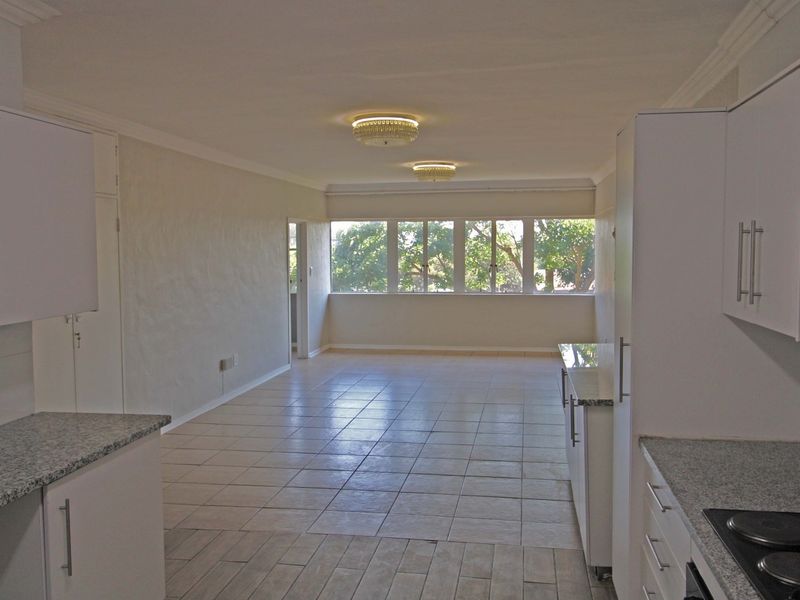 Spacious open-plan living and kitchen area featuring tiled flooring, modern white cabinetry with granite countertops, and large windows providing natural light.