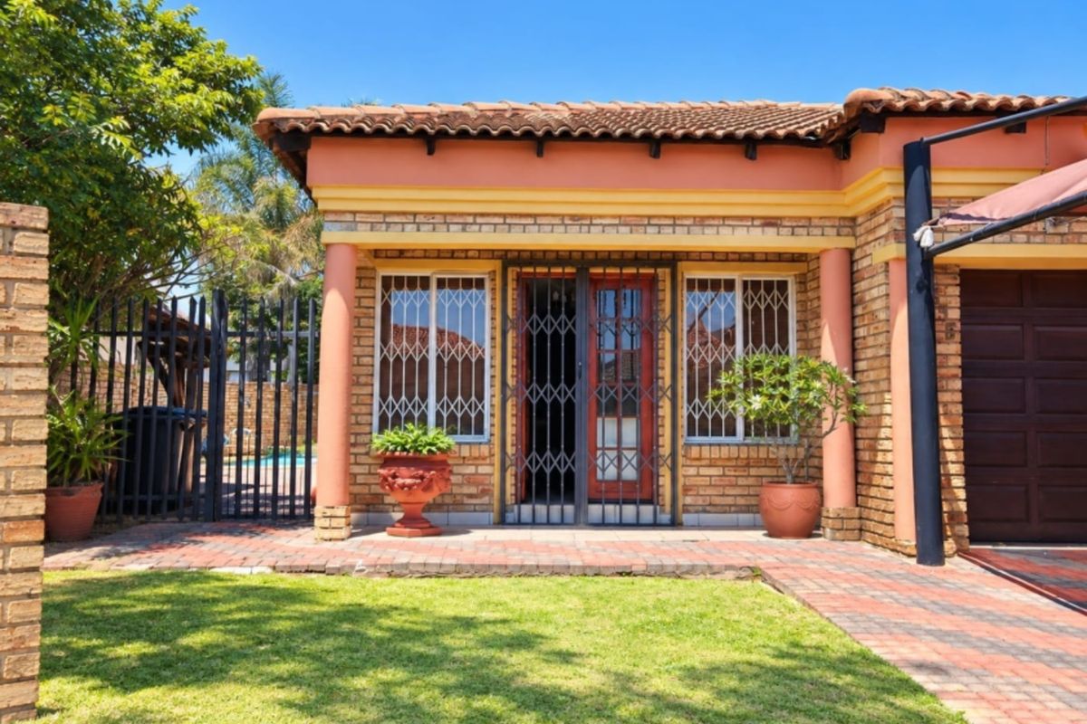 A well-maintained rear exterior view featuring a face brick facade, tiled roof, paved patio area, and secure metal fencing.