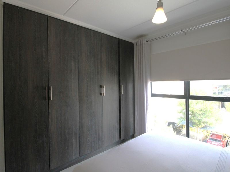 A well-lit bedroom featuring dark wood-finish built-in cupboards and a large window with roller blinds, offering a clean and functional living space.