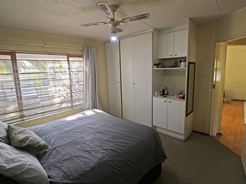 A bedroom featuring built-in cupboards, a ceiling fan, and large windows with venetian blinds, offering a functional space with potential for cosmetic updates.