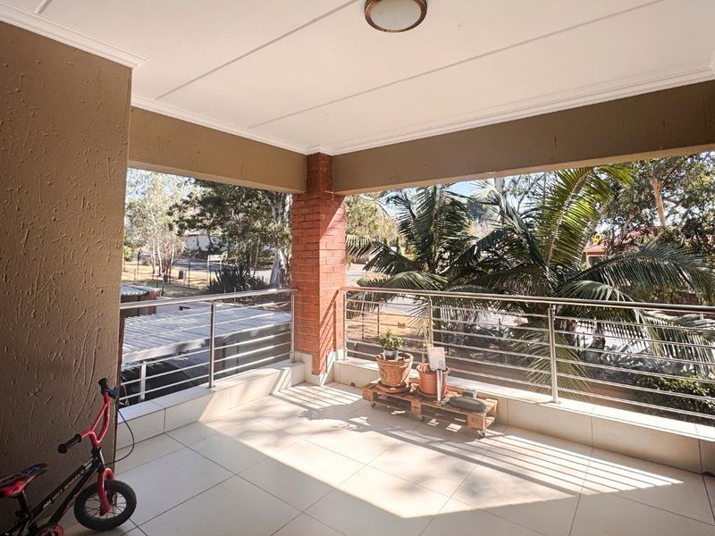 A spacious covered balcony featuring tiled flooring, a stainless steel balustrade, and an exposed brick pillar, offering an open outdoor living area.
