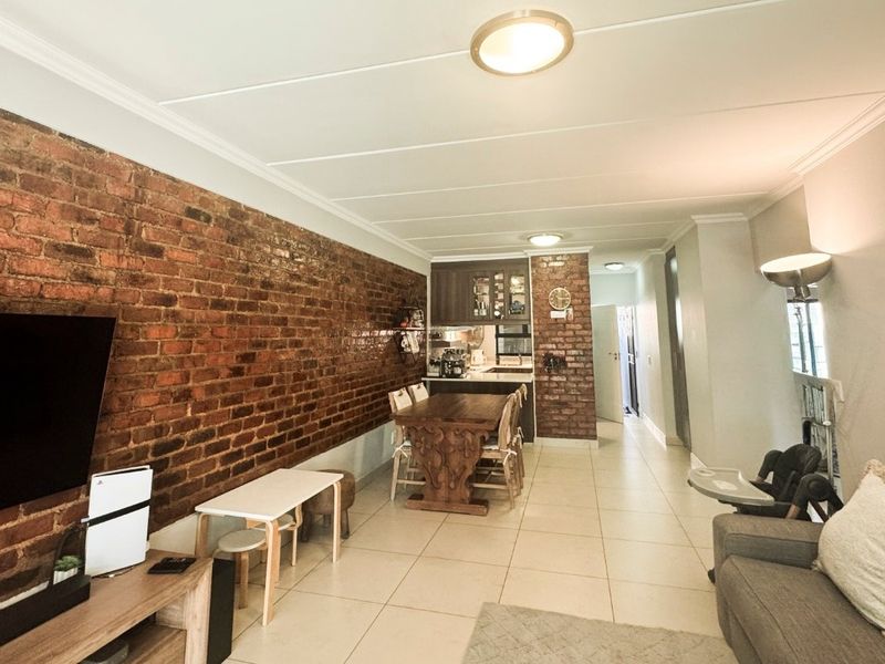 Open-plan living and dining area featuring a striking exposed brick feature wall, neutral tiled flooring, and integrated kitchen cabinetry.