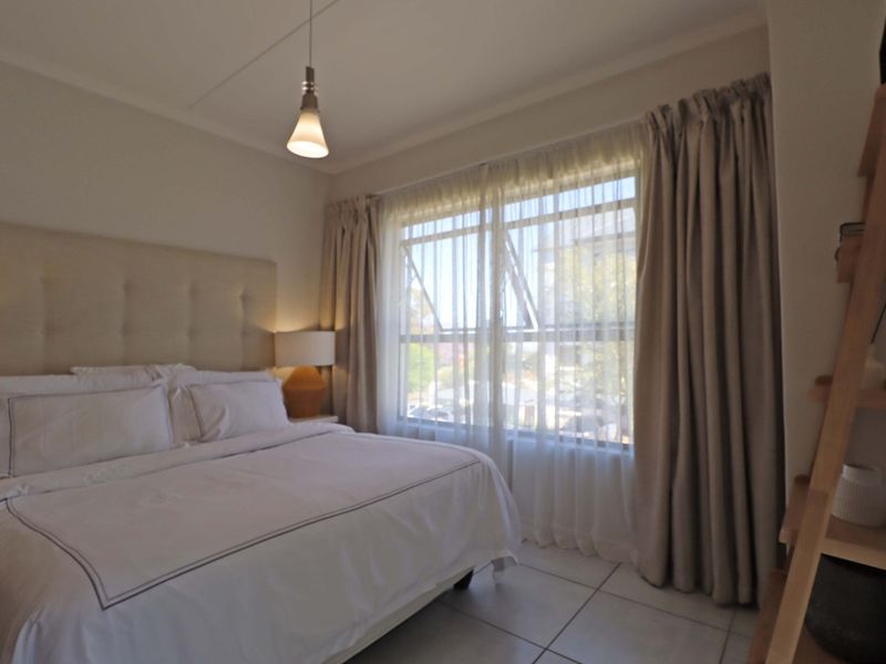 A well-maintained bedroom featuring neutral tiled flooring, a large window with curtain treatments, and modern pendant lighting.