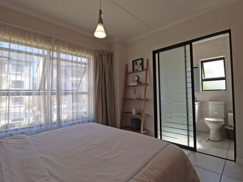 A well-lit bedroom featuring tiled flooring, a large window, and an en-suite bathroom accessed through a modern sliding glass door.