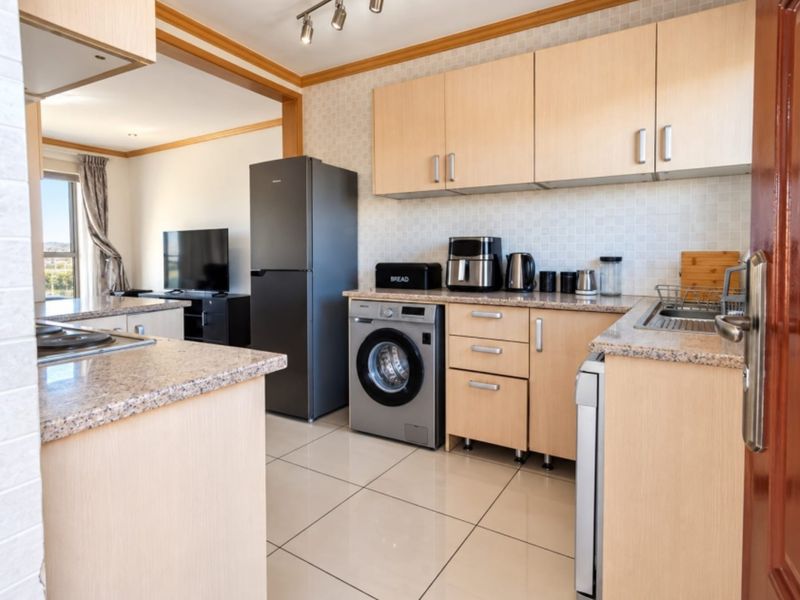 An open-plan kitchen featuring light wood cabinetry, granite countertops, tiled flooring, and integrated laundry facilities, flowing into a connected living area.