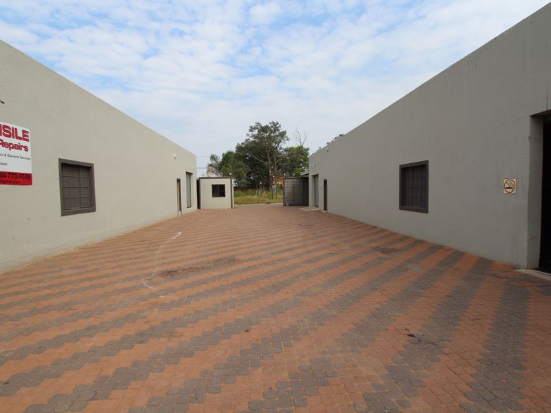 A paved commercial courtyard area situated between two industrial building wings, featuring security bars on windows and a small guard house at the rear.