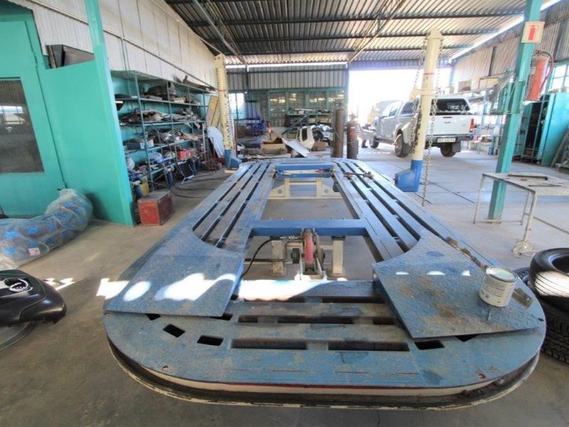 Industrial workshop space featuring a vehicle chassis straightening bench, metal storage shelving, and high-clearance corrugated roofing suitable for automotive repair operations.