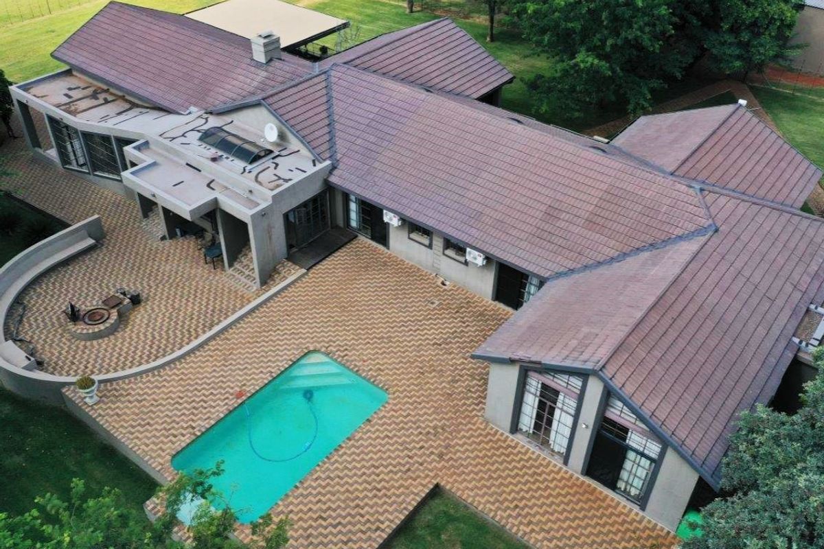 Aerial view of a farm property featuring a large tiled roof, swimming pool, paved patio area with a built-in fire pit, and surrounding garden.