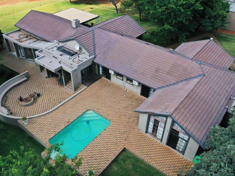 Aerial view of a farm property featuring a large tiled roof, swimming pool, paved patio area with a built-in fire pit, and surrounding garden.