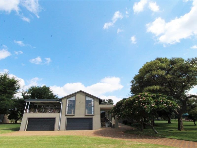Front view of a farm-style residence featuring a paved driveway, double garage, and an elevated covered balcony set within a spacious garden.