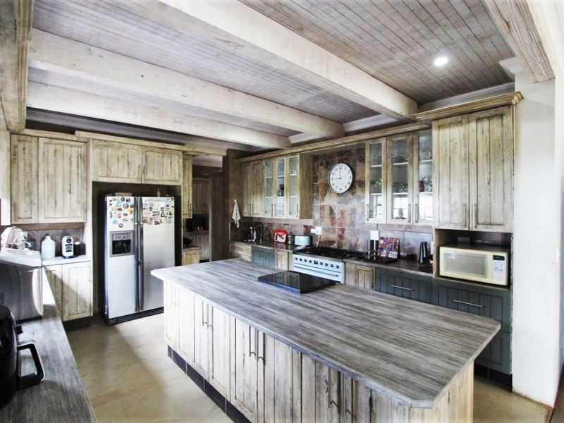 Spacious farmhouse kitchen featuring a large central island, rustic timber cabinetry, exposed ceiling beams, and stone tile backsplash with integrated appliances.