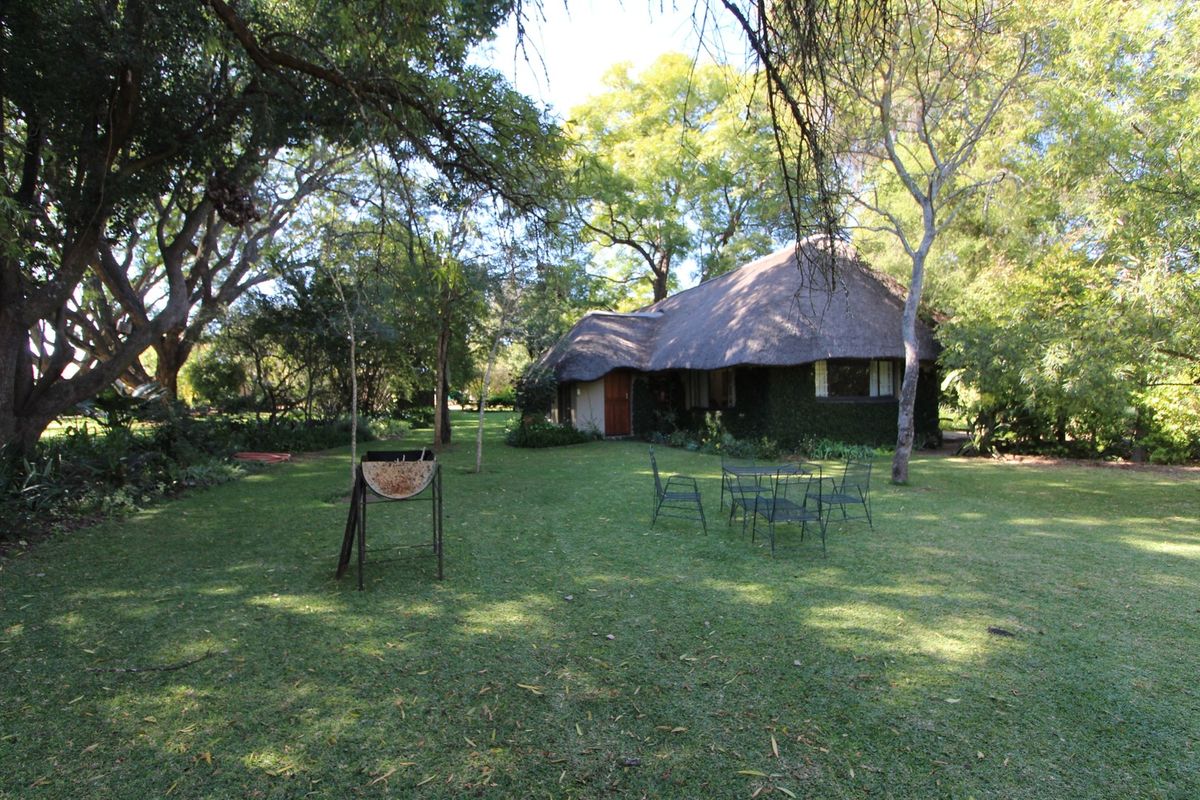 A spacious rear garden featuring a mature lawn, established trees, and a building with a traditional thatched roof.