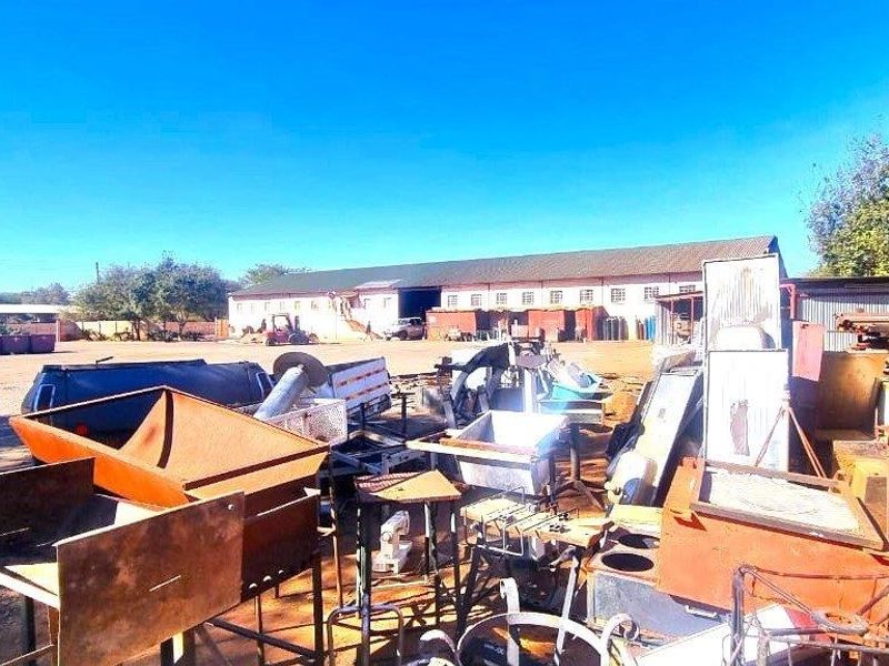 Expansive industrial yard area featuring a large warehouse structure, currently utilized for storage with significant accumulation of scrap materials and equipment.