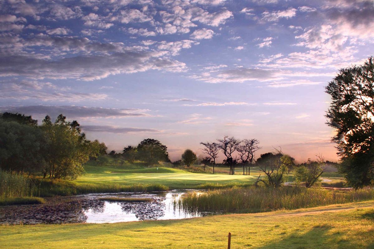 A scenic view of a well-maintained golf course fairway featuring a natural pond and mature trees under a sunset sky.