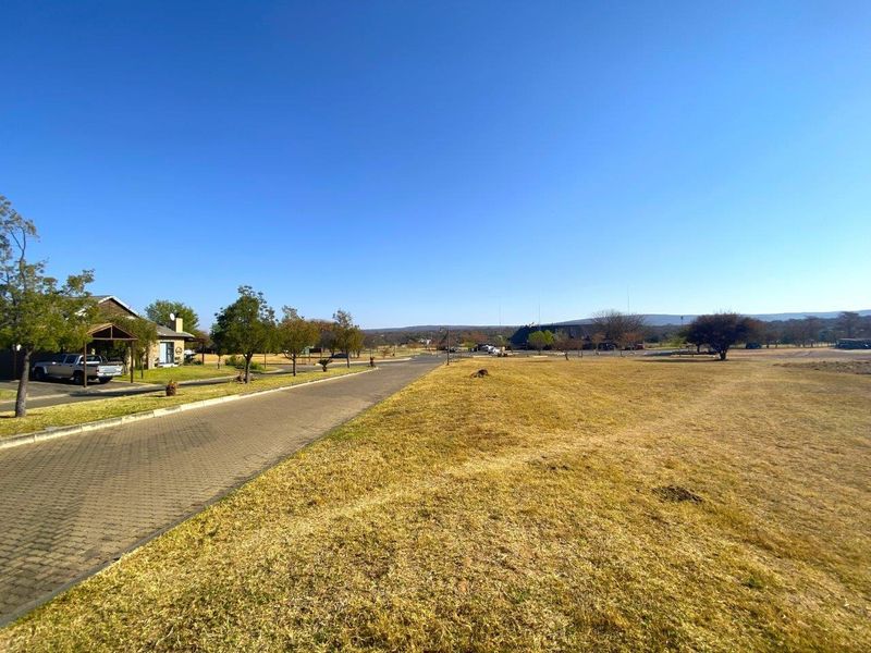 A wide-angle view of a vacant commercial land plot featuring a paved driveway, established verge, and open grass area under a clear blue sky.