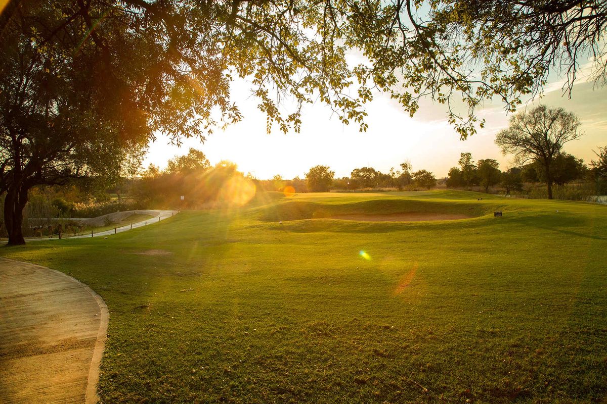 A scenic view of a well-maintained golf course featuring a sand bunker, paved pathways, and mature trees at sunset.