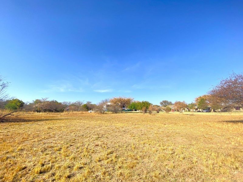 A wide-angle view of a vacant commercial land plot featuring dry grass and surrounding mature trees under a clear blue sky.