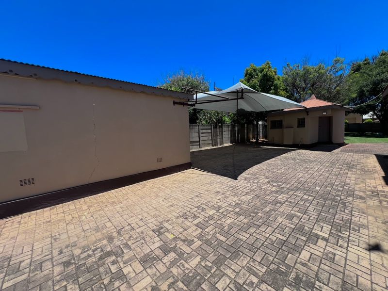 Rear exterior area featuring a paved driveway, a shade sail structure, and an outbuilding with a boundary wall.