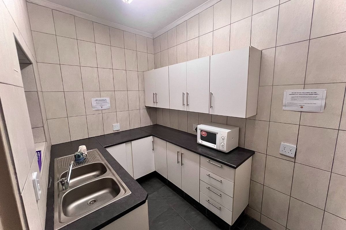 A functional office kitchenette featuring tiled walls, dark laminate countertops, double bowl stainless steel sink, and ample built-in white cabinetry.