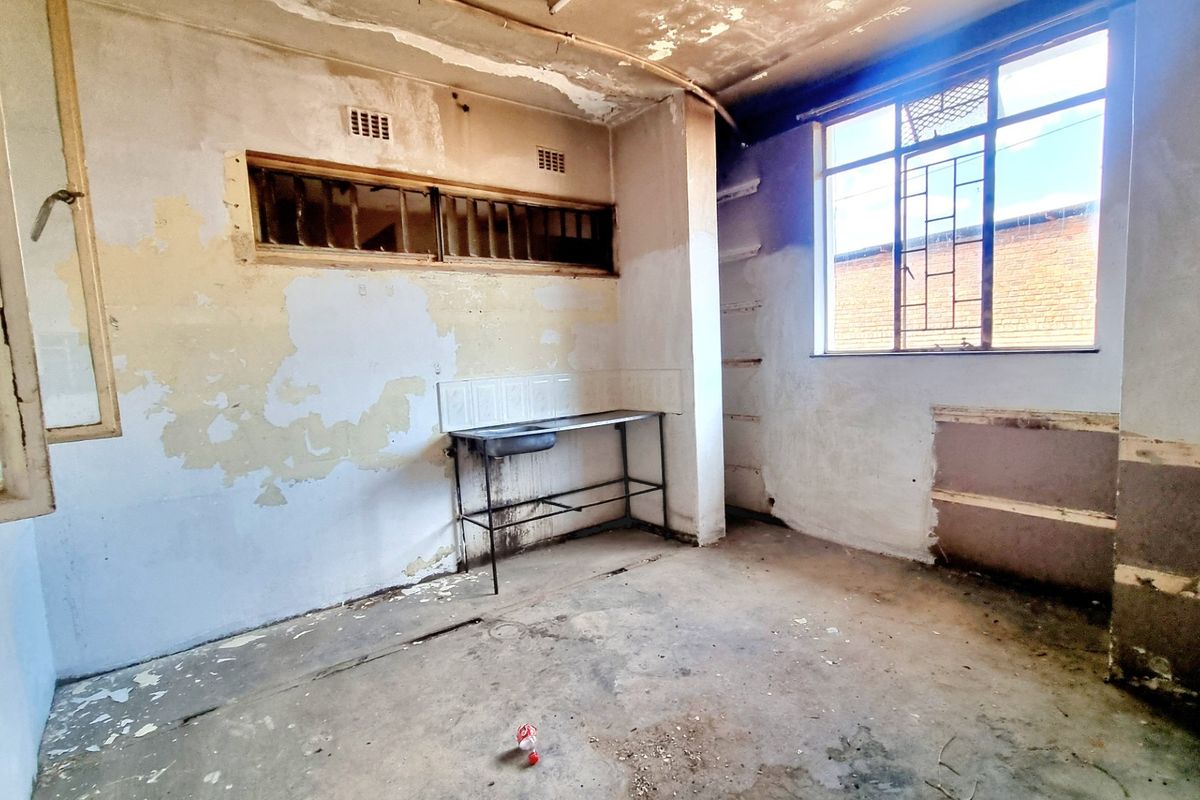 A vacant, dilapidated kitchen space requiring significant renovation, featuring a basic stainless steel sink, metal-framed windows with security bars, and damaged wall finishes.