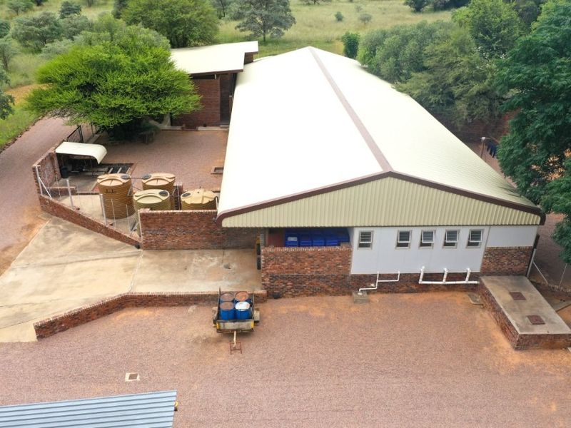 Aerial view of a farm building featuring a large metal roof, brick exterior walls, and external water storage tanks on a gravel property.