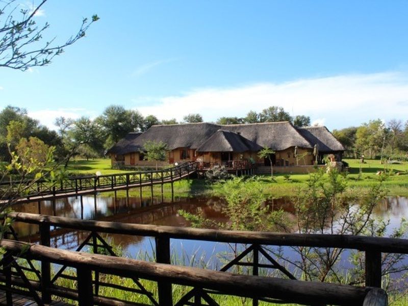 A scenic view of a thatched-roof farm property featuring a wooden footbridge crossing a tranquil pond, surrounded by lush green gardens.