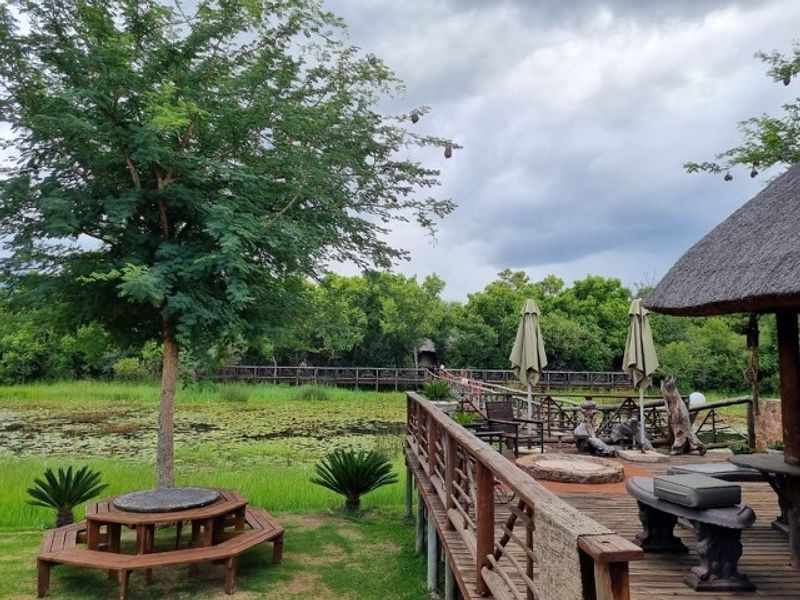 A scenic outdoor wooden deck featuring a thatched roof structure, overlooking a tranquil pond and lush green landscape with a raised wooden walkway.