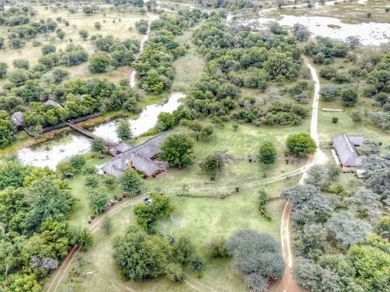Aerial view of a rural farm property featuring multiple thatched-roof structures, a natural pond, a footbridge, and surrounding bushveld landscape.