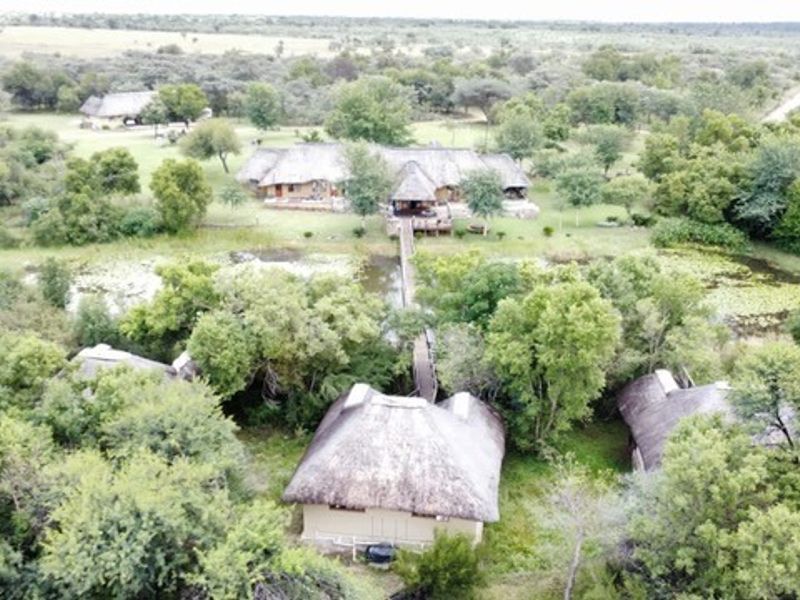 Aerial view of a farm property featuring multiple structures with thatched roofs, connected by wooden walkways over a natural pond and lush surroundings.