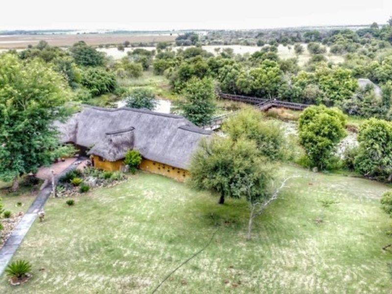 Aerial view of a farm property featuring a traditional thatched-roof building, expansive green lawns, mature trees, and a wooden boardwalk over a natural area.