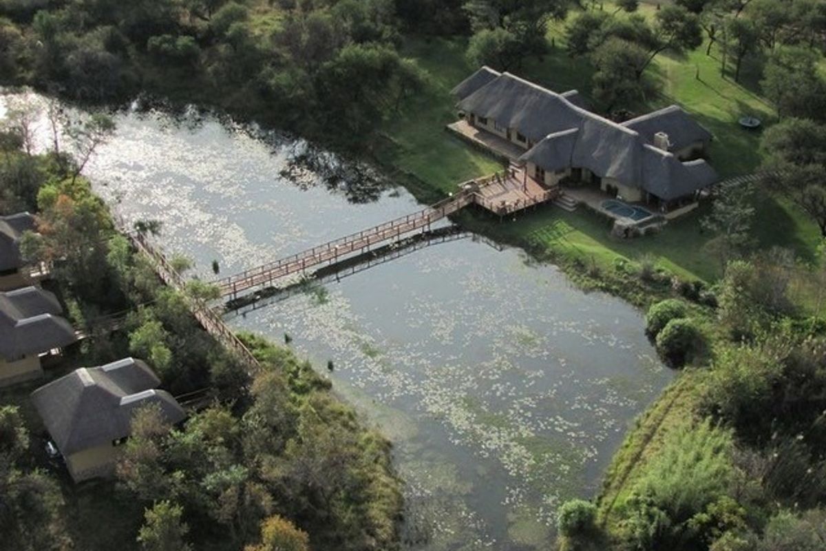 Aerial view of a farm property featuring a thatched-roof main house, a swimming pool, and a wooden footbridge crossing a scenic pond.