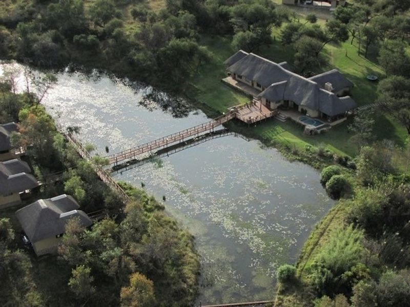 Aerial view of a farm property featuring a thatched-roof main house, a swimming pool, and a wooden footbridge crossing a scenic pond.
