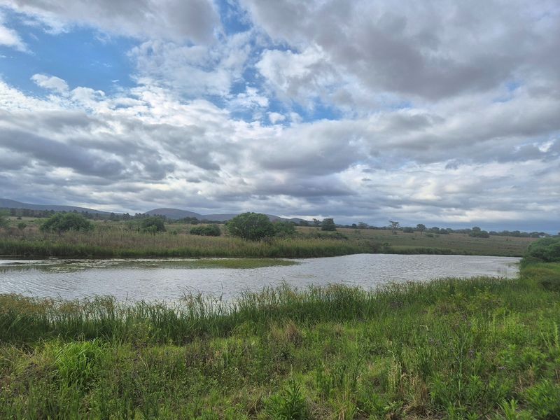 A scenic view of a natural dam surrounded by lush green grassland and rolling hills under a cloudy sky.