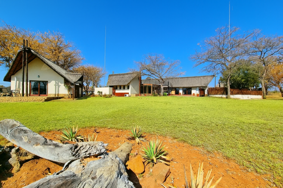 Spacious rear garden view featuring multiple structures with traditional thatch roofing, stone accents, and expansive lawn area under a clear blue sky.