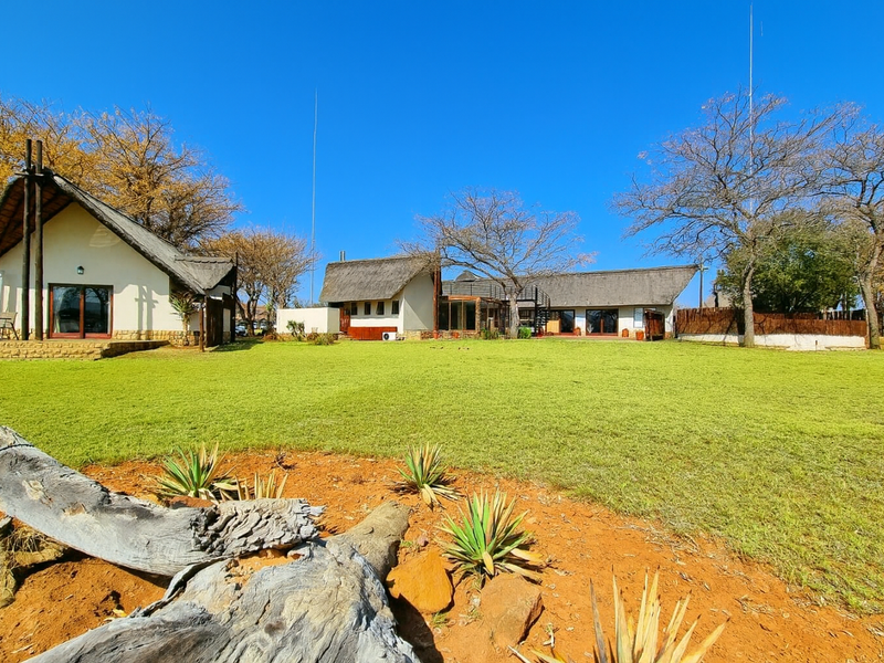 Spacious rear garden view featuring multiple structures with traditional thatch roofing, stone accents, and expansive lawn area under a clear blue sky.